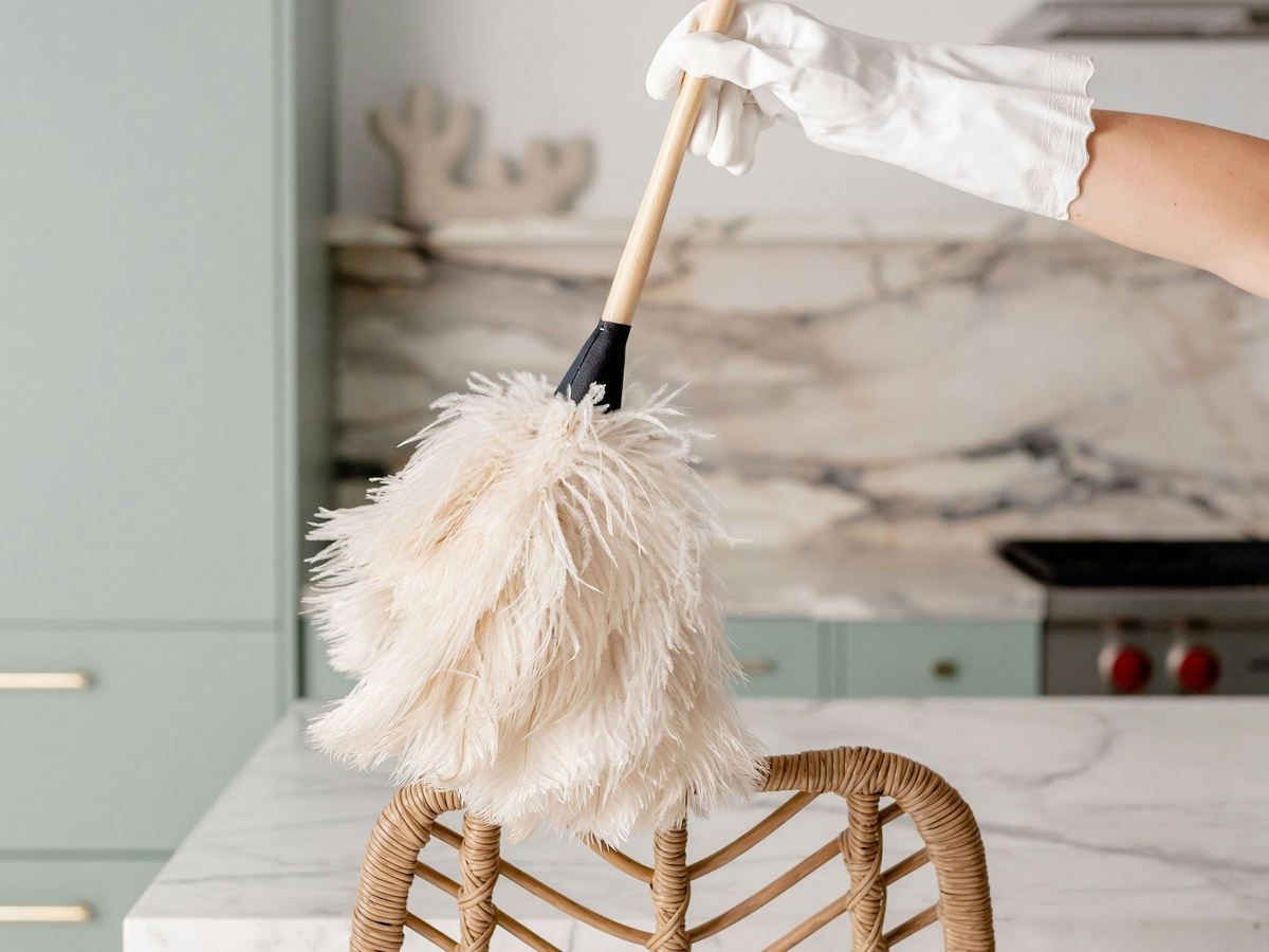 A gloved hand dusting a rattan chair with a feather duster in a bright kitchen